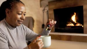 Cbd anxiety treatment - African senior woman taking cannabis oil in tea cup by cozy fireplace at home