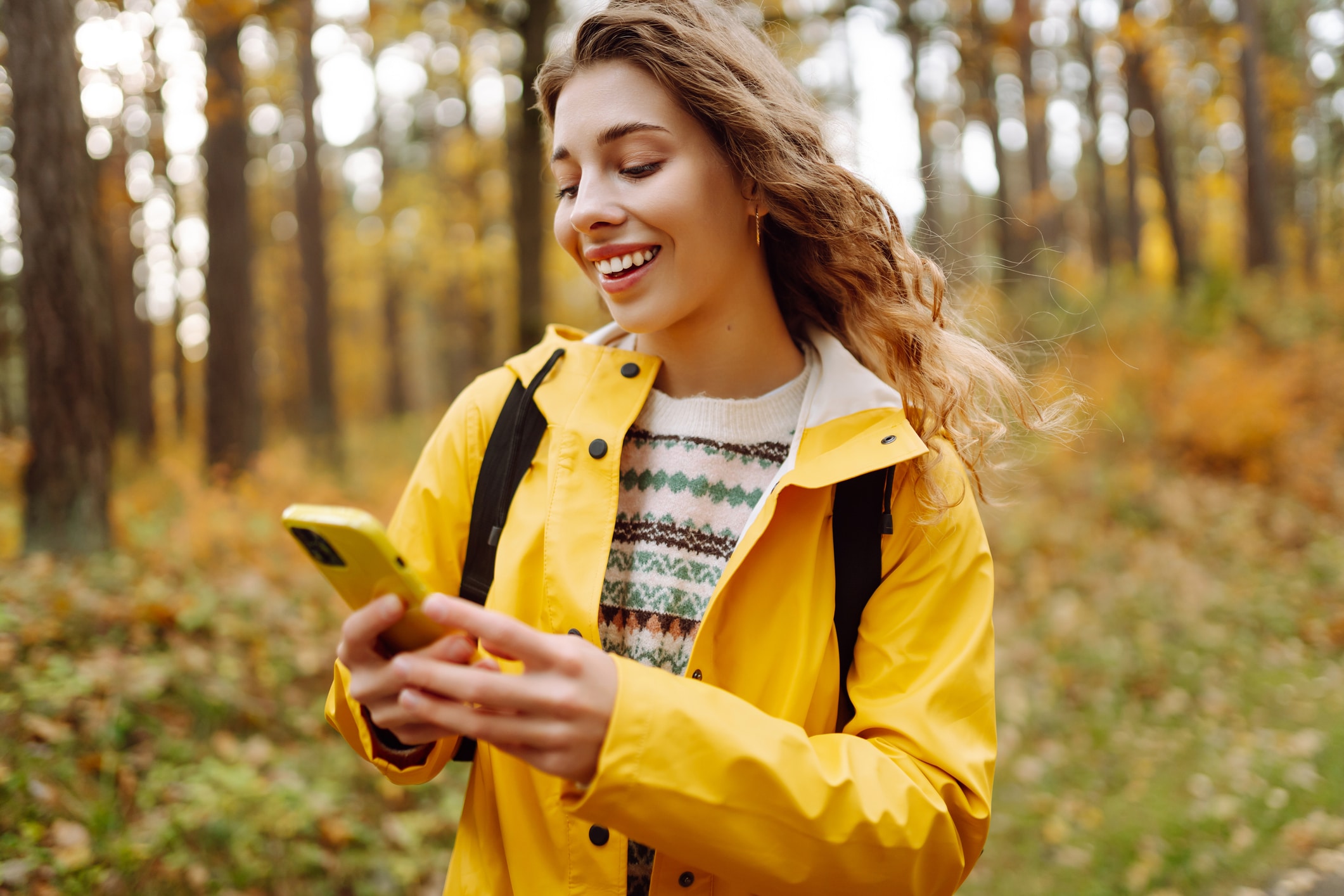 Happy woman in yellow raincoat and with phone on path in sunny autumn park. Young woman blogging or chatting while relaxing outdoors. Walking, blogging concept.